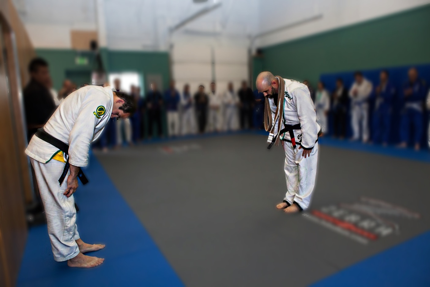 Two Brazilian Jiu-Jitsu practitioners in white gis bowing on the mat during a ceremony at Busy BJJ, students watching respectfully in the background